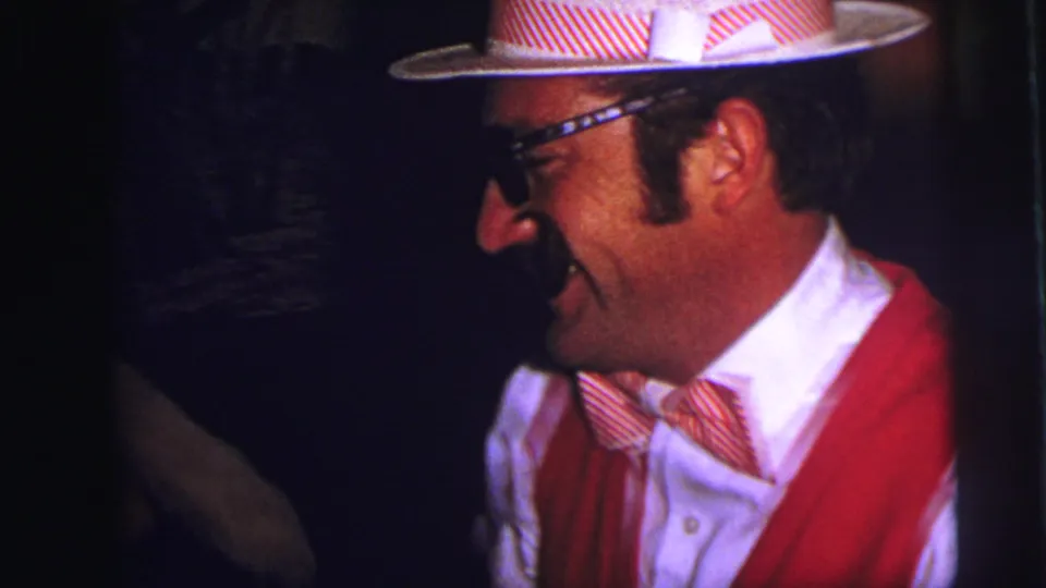 In 1973 Lynbrook home-movie footage, a costumed man in a red vest, striped bow tie, glasses, and hat smiles inside a dim party room.