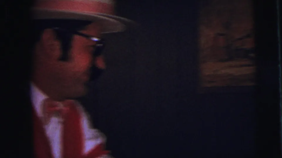 A close, dark 1973 Lynbrook party frame catches the costumed man partly off-center, with his hat, glasses, bow tie, and red vest visible.