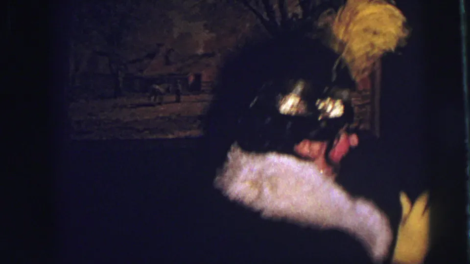 In the 1973 Lynbrook party footage, a second costumed adult appears in a feathered hat and white fur-trimmed wrap inside the dim room.