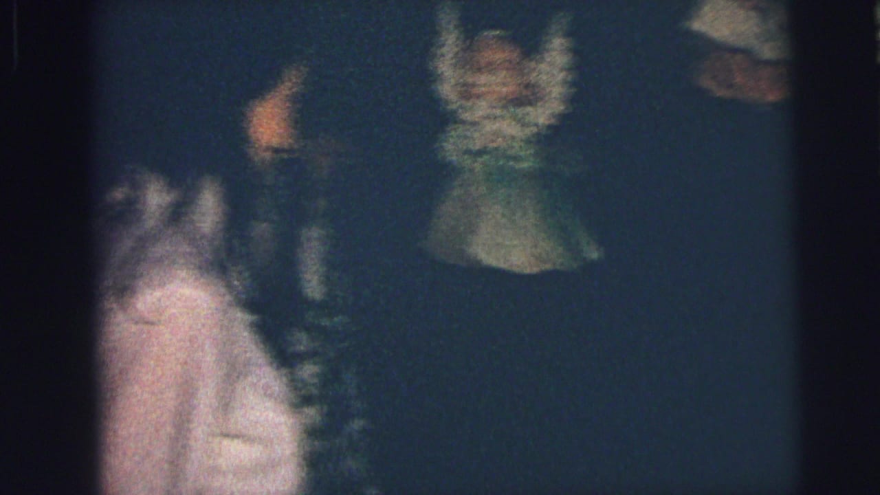1973 Lynbrook home-movie frame showing figures in a dim interior beside a Christmas tree and pale patterned curtains.