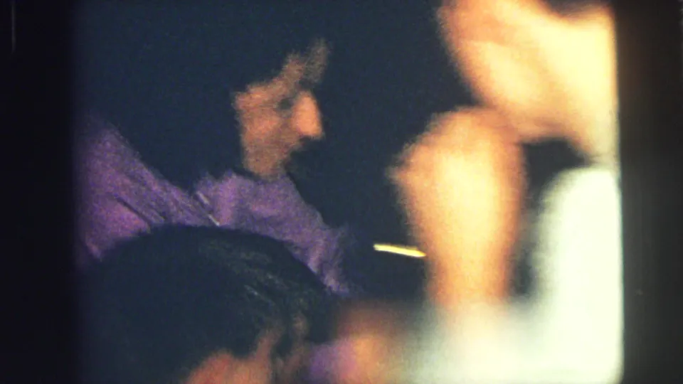 Grainy Lynbrook home-movie frame with a seated person in purple clothing and blurred movement in the foreground.