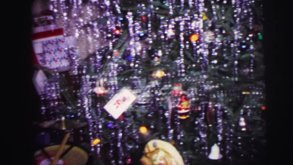 1973 Lynbrook home-movie frame centered on a tinseled Christmas tree with lights, ornaments, a gift tag, and toys beneath.
