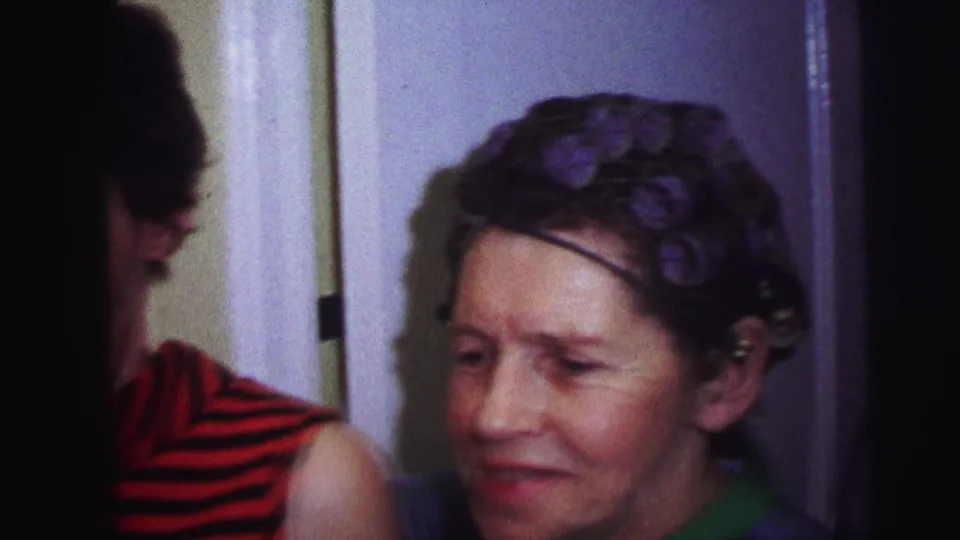 1973 Lynbrook home-movie frame centered on an older woman in hair rollers, with the younger woman partly cropped at the left edge.