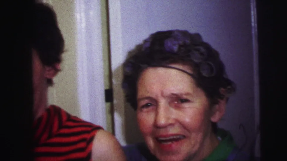 1973 Lynbrook 8mm frame of an older woman in hair rollers facing forward, with a younger woman in a striped top partly visible.
