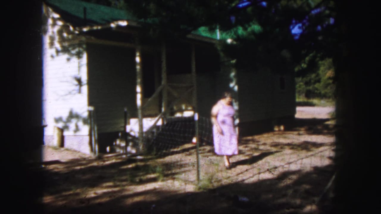 Woman in a purple dress walks away from a white house across a shaded dirt yard.