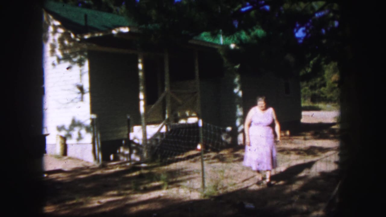 A woman in a purple dress approaches the camera from the yard outside a white house in Colorado Springs, 1958.