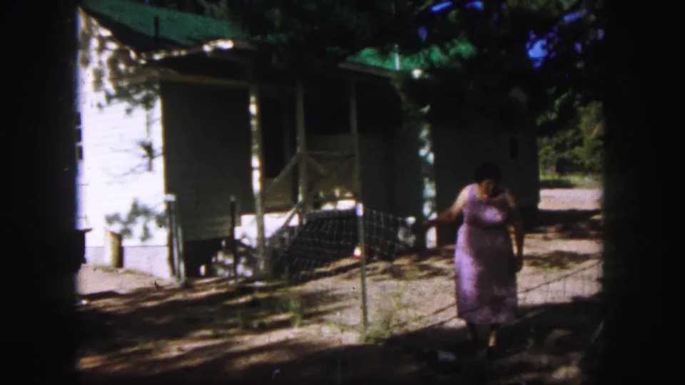 1958 Colorado Springs home-movie frame showing a woman in a purple dress approaching a modest white house through a shaded yard.