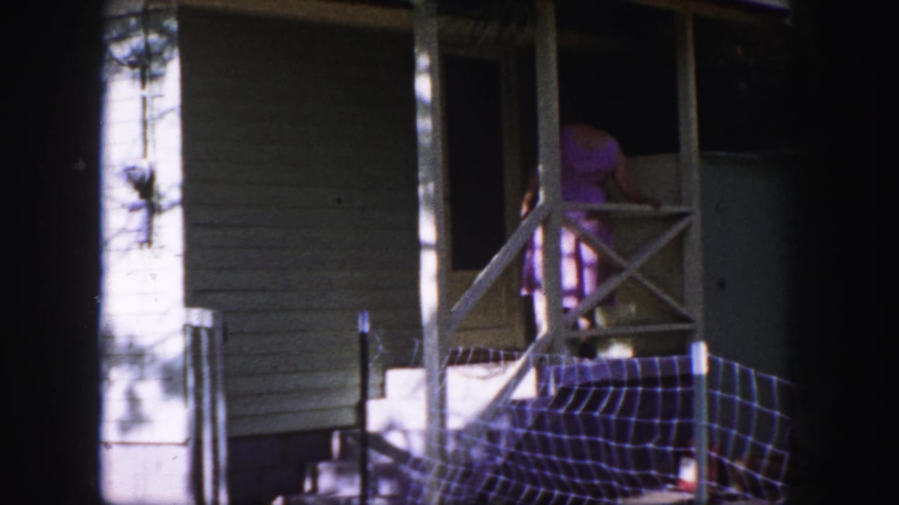 Woman in pink skirt stands at a porch screen door beside white railings and shaded siding.