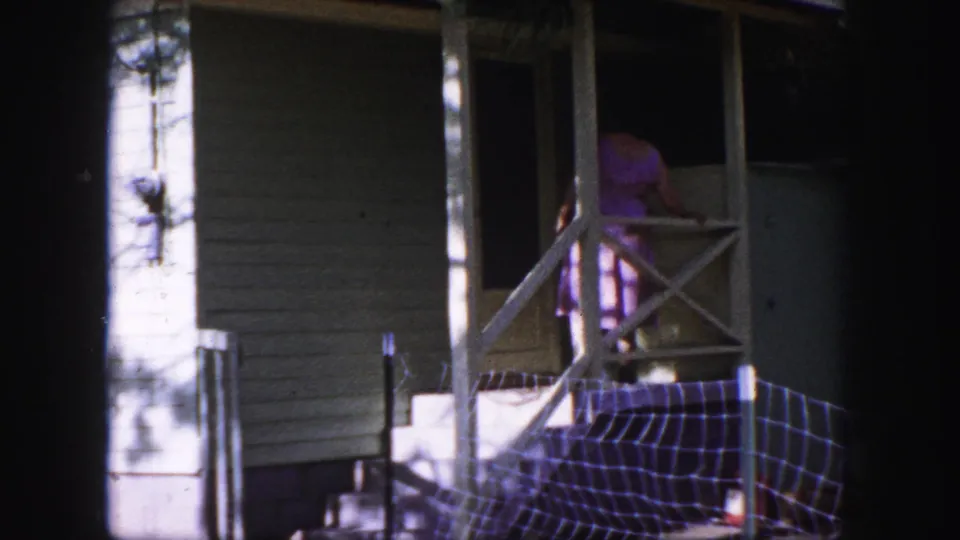 In 1958 Colorado Springs home-movie footage, a woman in a pink skirt stands on a porch near a screen door and crossed railing.