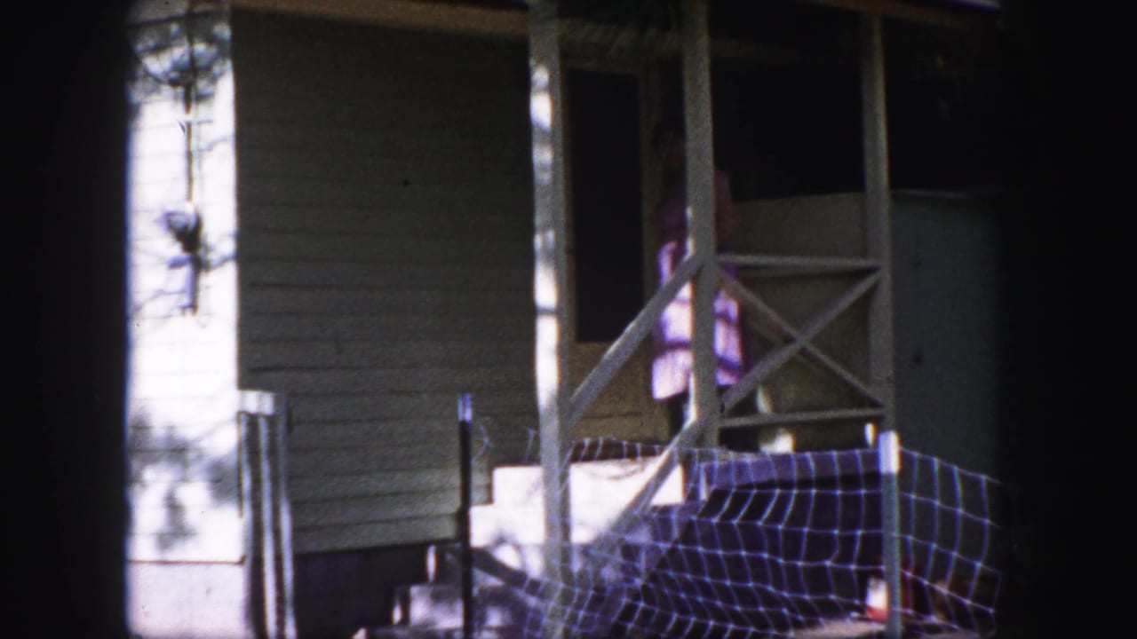 Woman reaches toward or opens the porch screen door beside a white railing and shaded wall.