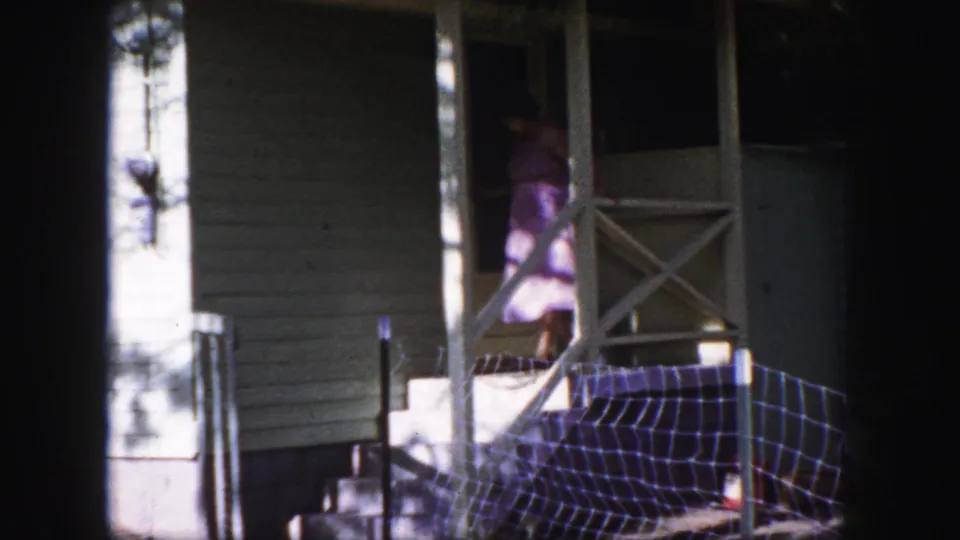 The woman moves through the porch doorway into the house, framed by crossed railing, siding, and bright sun patches.
