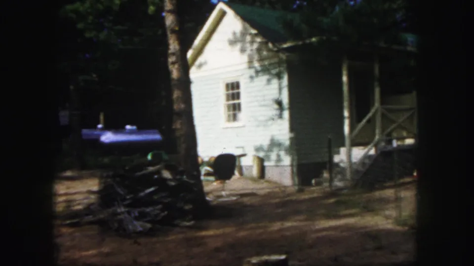 1958 Colorado Springs home-movie frame showing the side of a house, porch area, window, yard, and stacked outdoor items.