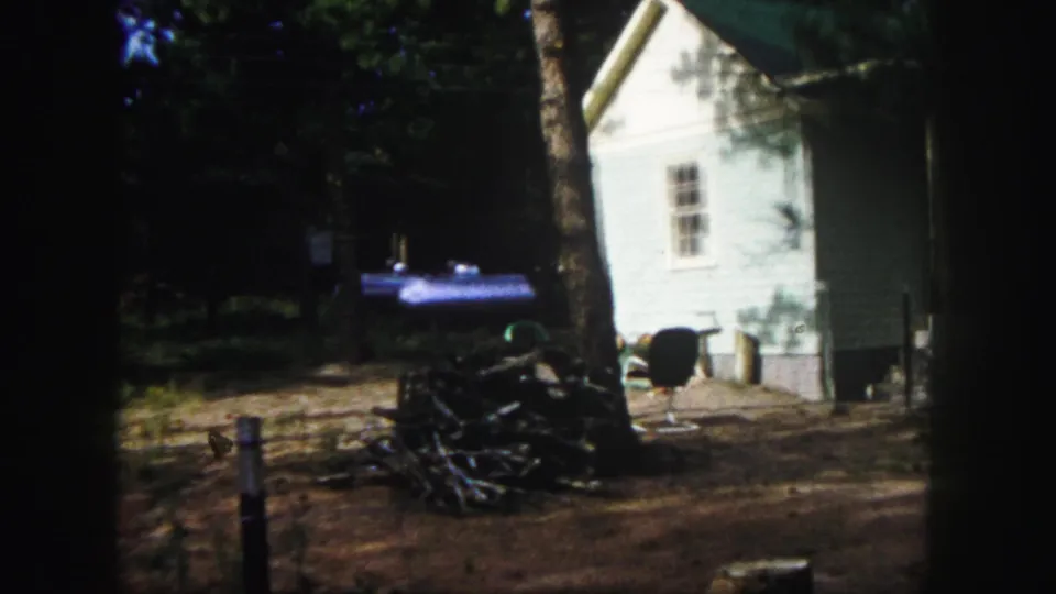 1958 Colorado Springs frame showing the side wall of a modest house with yard objects, tree shade, and the porch edge nearby.