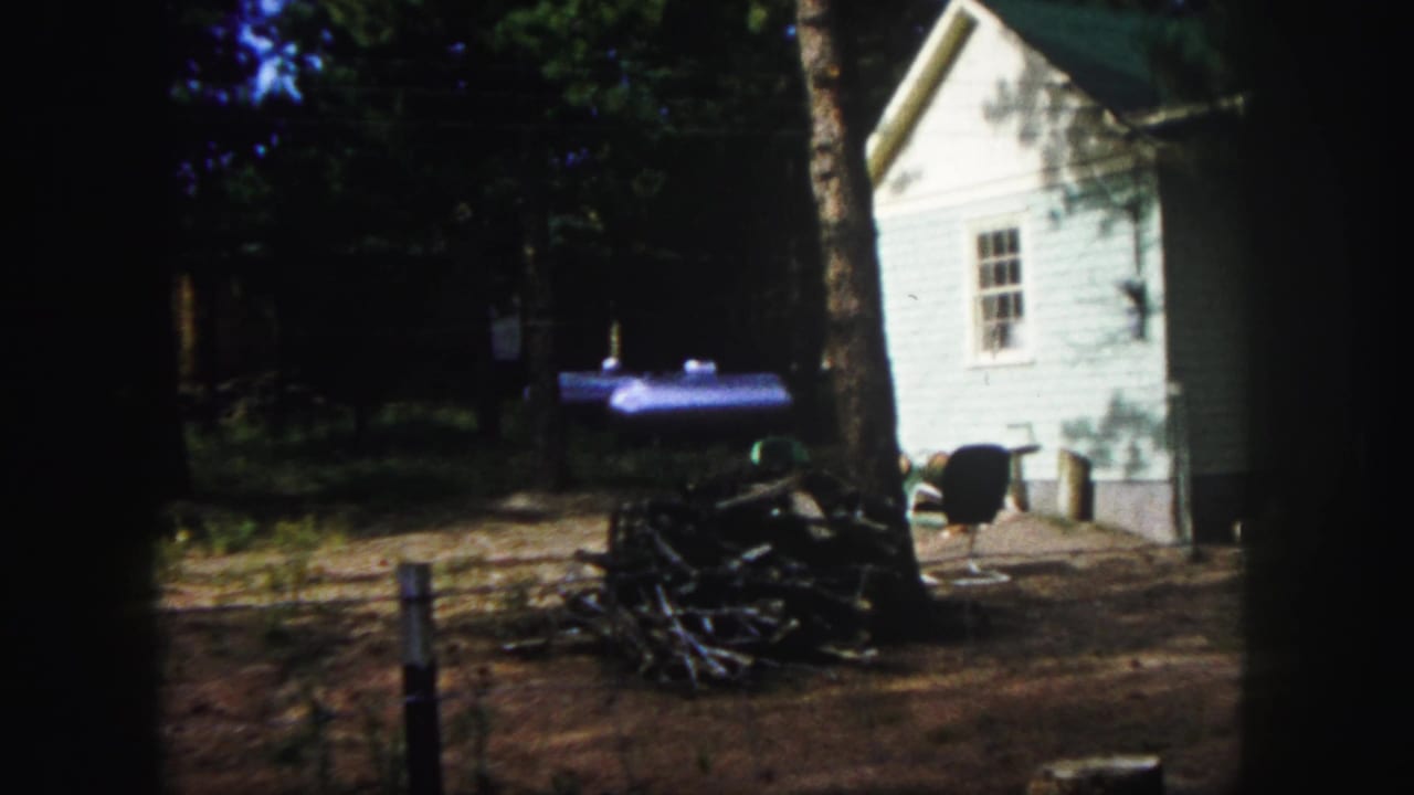 White clapboard building beside trees, with stacked firewood and a dark chair in a dirt yard.
