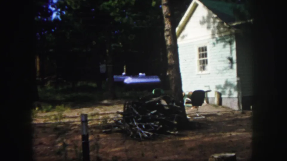 1958 Colorado Springs home-movie frame showing a white clapboard building partly shaded by trees, with firewood and outdoor seating in the yard.