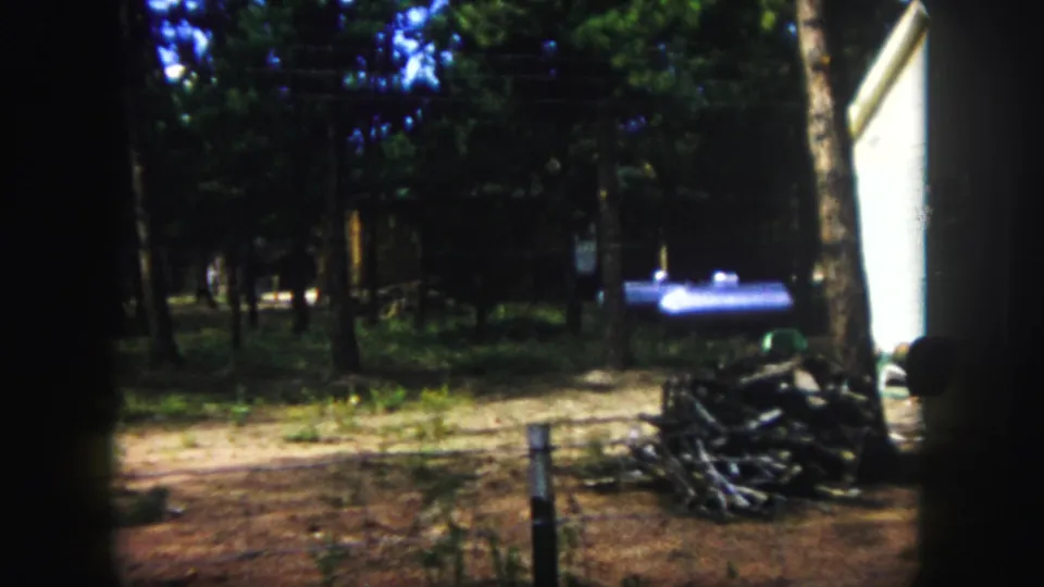 1958 Colorado Springs home-movie frame looking across a wooded dirt yard toward a white building edge, firewood, chair, and blue object.
