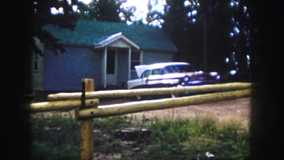 1958 Colorado Springs frame showing a small blue house among trees, a split-rail fence, and a parked light-colored 1950s station wagon.