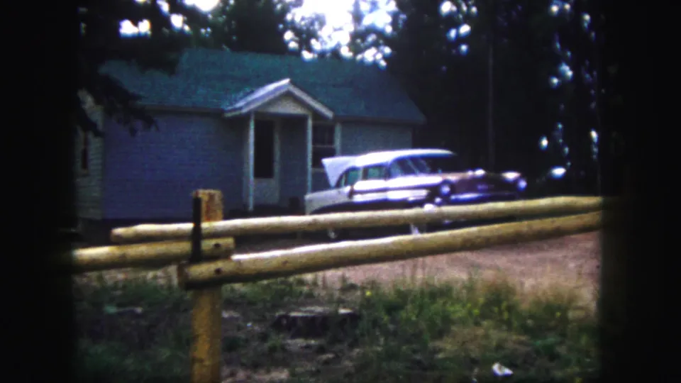 1958 Colorado Springs home-movie view of a small house, split-rail fence, dirt driveway, and parked 1950s station wagon among trees.