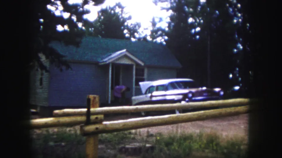 1958 Colorado Springs home-movie frame showing a woman in pink approaching a grey house beside a white and purple station wagon.