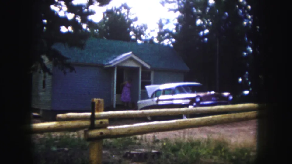 1958 Colorado Springs frame with a woman in pink at the doorway of a grey house, with a vintage station wagon parked outside.