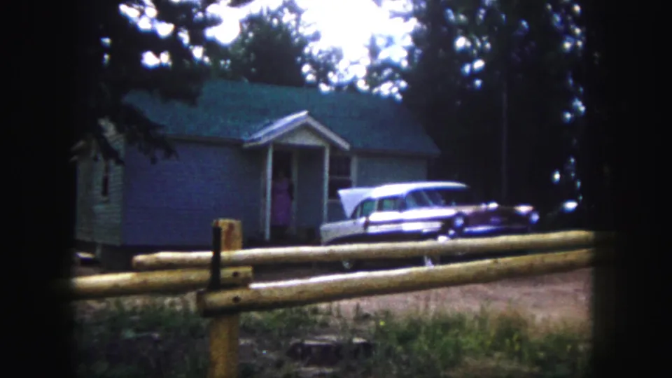 1958 Colorado Springs home-movie frame showing the woman at the doorway, with the car and split-rail fence still in view.