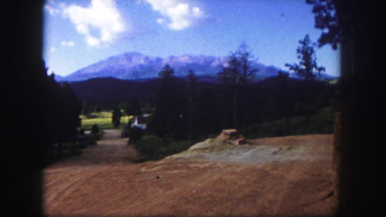 Dirt path and open clearing framed by dark trees with distant mountains under a blue sky.
