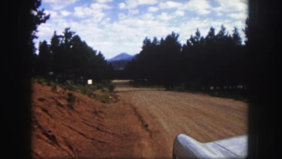 1958 Colorado Springs road-view frame with a winding dirt road, pine trees, distant peak, and white car fender.