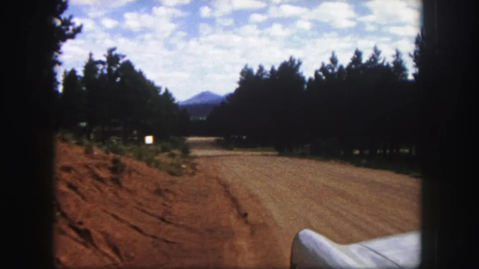 1958 Colorado Springs home-movie view down a dirt road bordered by pine trees and a low mountain peak.