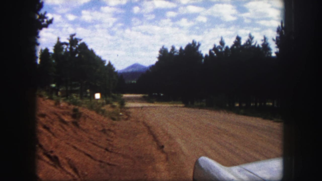1958 Colorado Springs home-movie frame showing a dirt road between pines, a distant mountain, cloudy sky, and a white car fender.