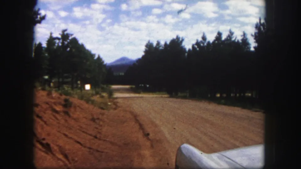 1958 Colorado Springs road scene from inside a moving car, with a curving dirt road, pine forest, mountain view, and car fender.