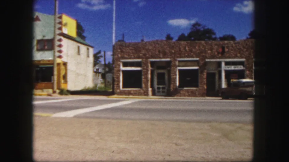1958 Colorado Springs street view showing a stone storefront, adjacent striped building, crosswalk markings, and a parked mid-century car.