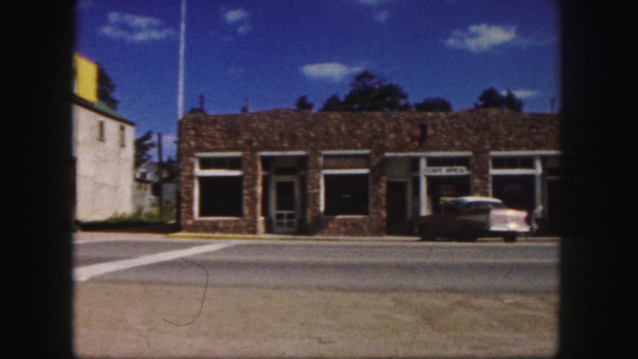 1958 Colorado Springs street view with low stone storefronts, a cafe sign, and a parked mid-century sedan across a broad road.