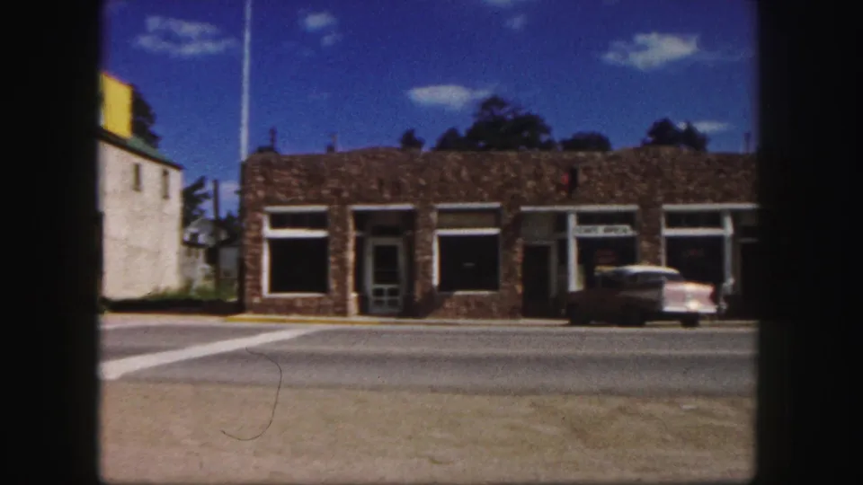 1958 Colorado Springs street view with low stone storefronts, a cafe sign, and a parked mid-century sedan across a broad road.