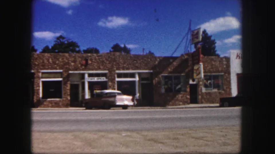 1958 Colorado Springs street pan with the cafe storefront, parked sedan, and part of a neighboring business sign coming into view.