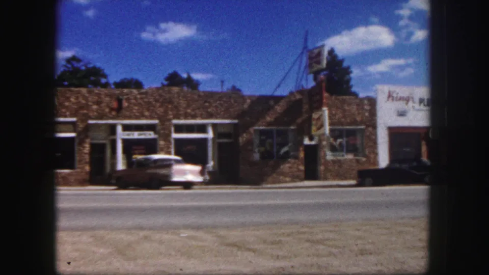 1958 Colorado Springs storefront row with a cafe, King's Plumbing sign, parked sedan, and another car near the curb.