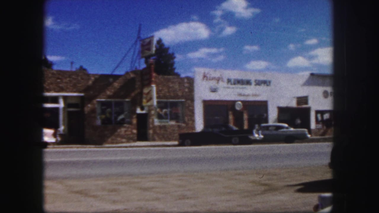 Storefront row with King's Plumbing Supply, parked cars, and dark film border.
