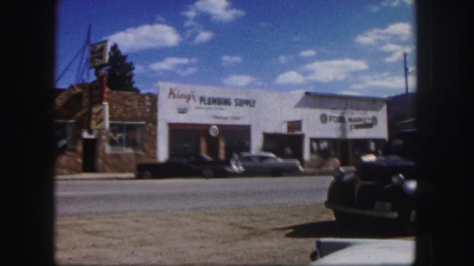 1958 street view of King's Plumbing Supply, adjacent signage, parked sedans, and a dark car partly in the foreground.