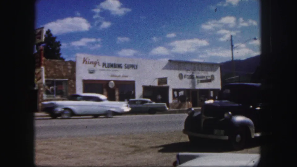 A light-colored 1950s car passes King's Plumbing Supply in a sunny Colorado Springs street scene.