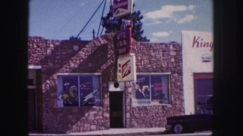 1958 Colorado Springs street-front frame showing stone storefronts, beer advertising panels, a vertical sign, and a dark parked car.