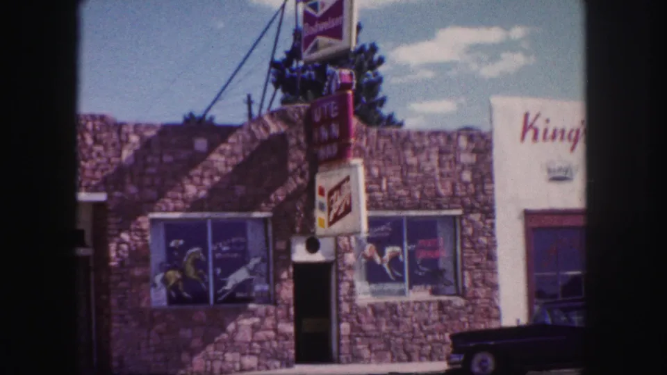 1958 Colorado Springs exterior view of a stone-faced storefront with beer signs, business lettering, and a dark car parked along the street.