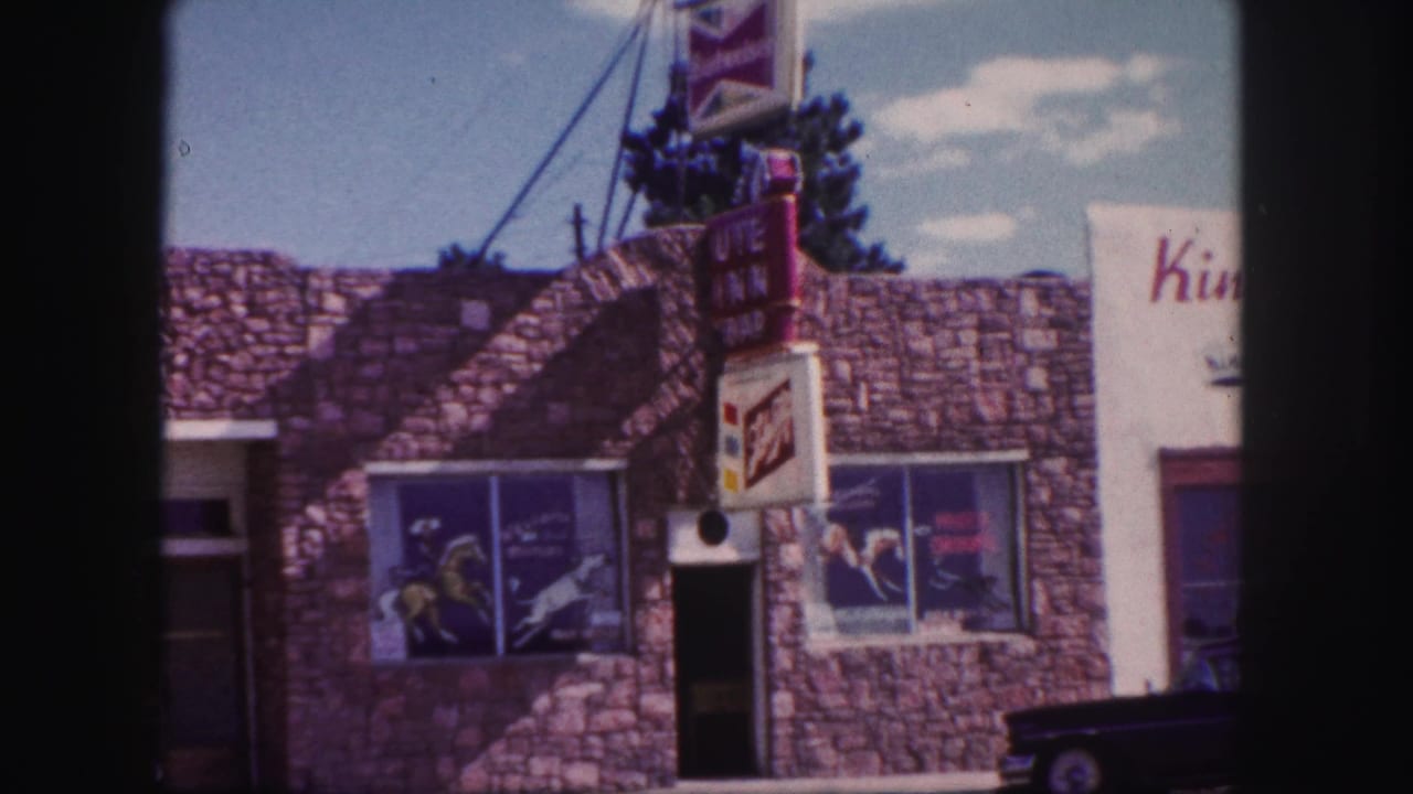 1958 Colorado Springs home-movie frame showing a stone-faced roadside motel with commercial signs and display windows.