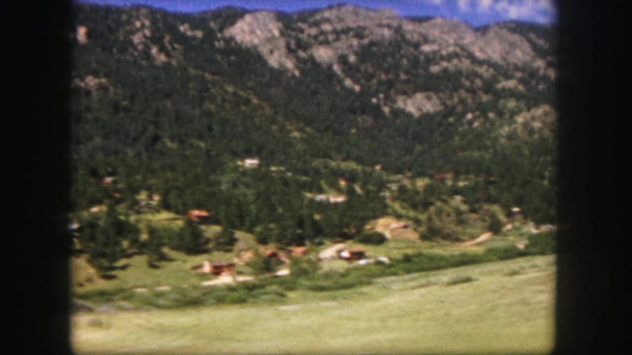 1958 Colorado-area home-movie view of a green valley, small structures, dense pine slopes, and rocky mountains beyond.