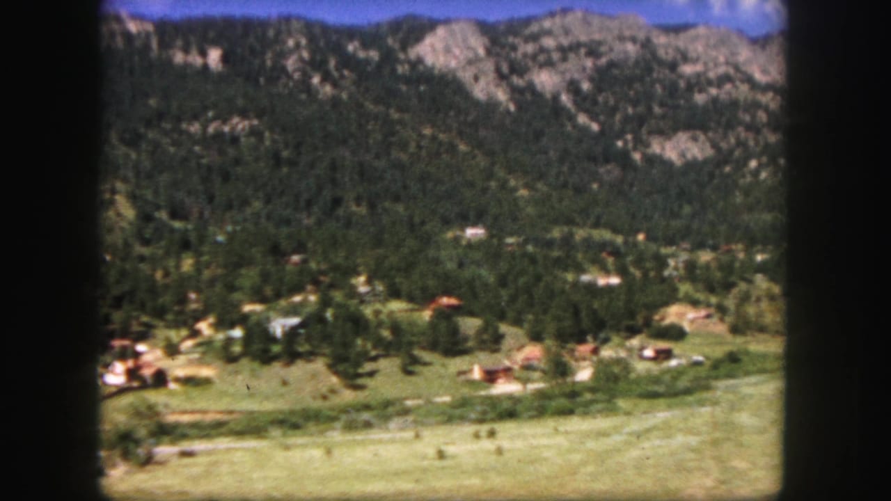 Mountain valley with small buildings, trees, open grass, and rocky slopes in the distance.