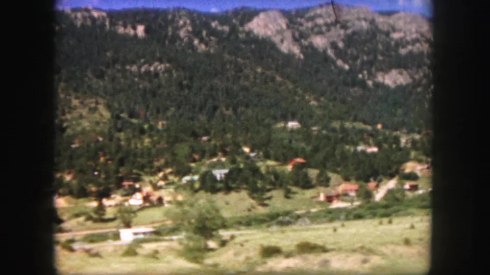 1958 home-movie frame from a moving vehicle showing a Colorado mountain valley with scattered structures and forested ridges.