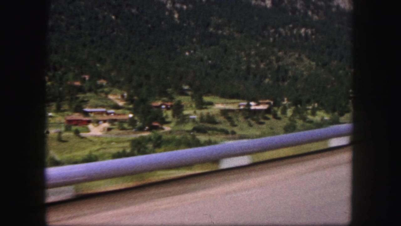 1958 Colorado Springs home-movie view from a moving vehicle past a railing toward cabins, open grass, and a forested slope.