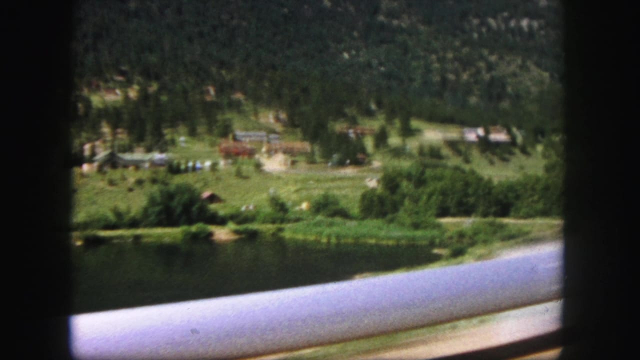 Lake edge, green field, scattered buildings, pine trees, and a pale railing passing close to camera.