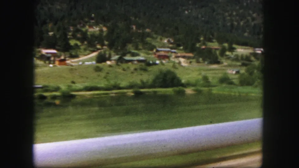 Passenger-side 1958 Colorado Springs view with motion blur across a lake, grassy bank, distant cabins, and pine-covered terrain.