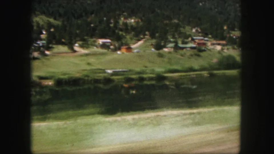 1958 Colorado Springs home-movie frame showing a lakeside field, reflected tree line, distant buildings, and vehicle-window framing.