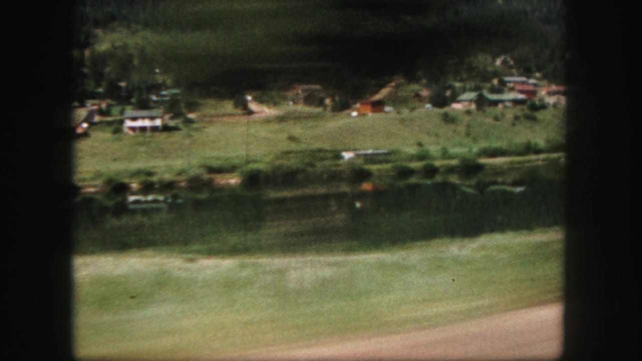 1958 Colorado Springs-area home-movie view from a moving car passing a rural settlement near water or open ground and wooded hills.