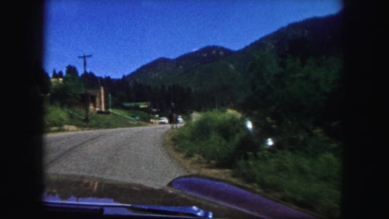 Paved road curves through green hills with a car hood visible at the bottom of the frame.